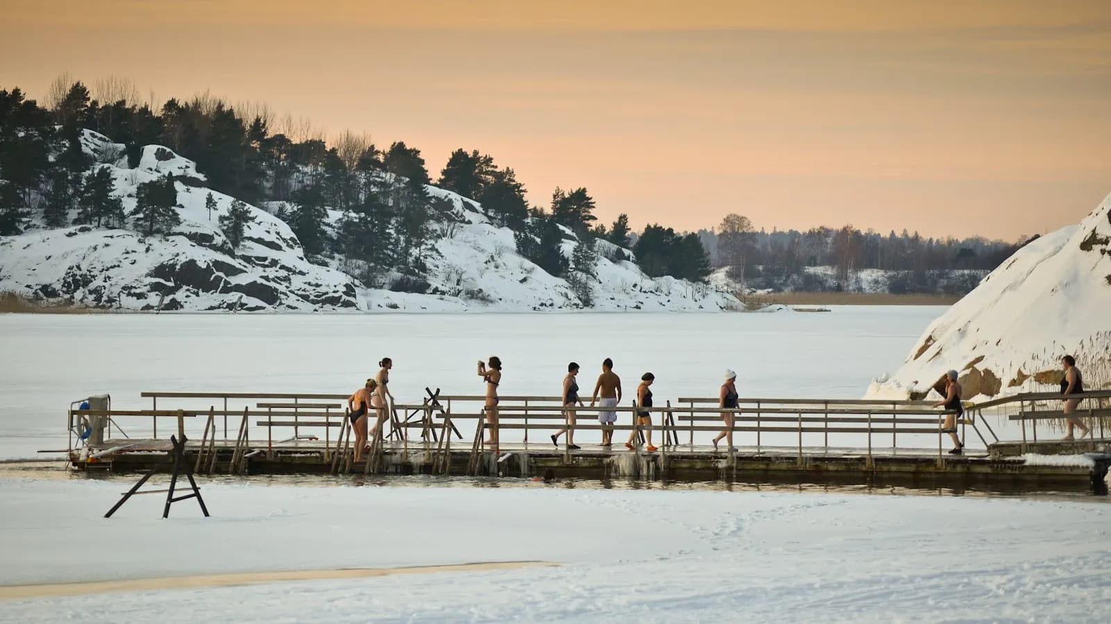 Winter swimmers in swimsuits on a wooden pier beside a hole cut in the sea ice at Turku, Finland, in low afternoon sun