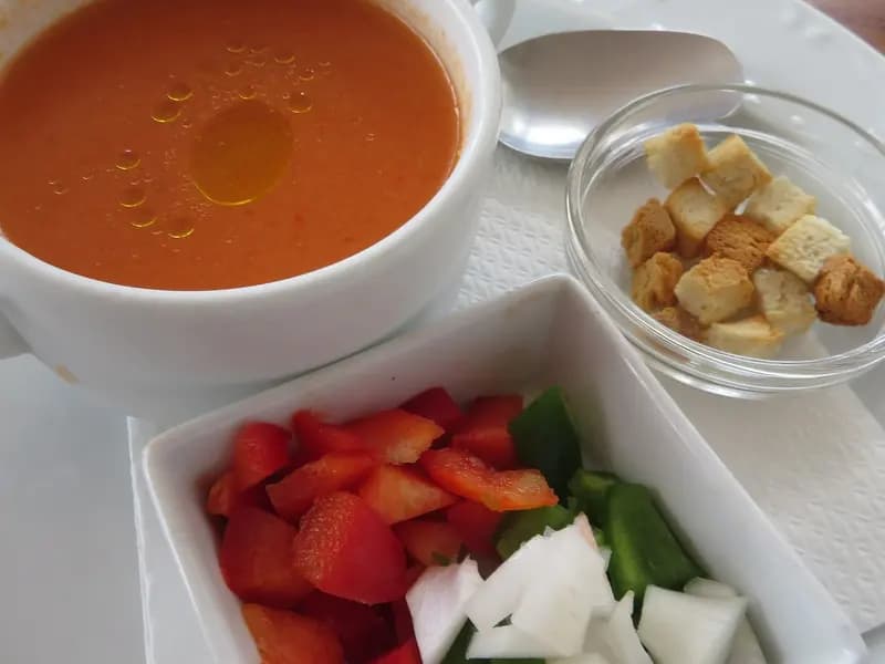 A cup of gazpacho andaluz with a thread of olive oil on top, beside small dishes of diced pepper, cucumber, onion and croutons for garnish