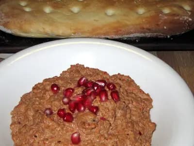 A plate of muhammara scattered with pomegranate seeds, with flatbread behind