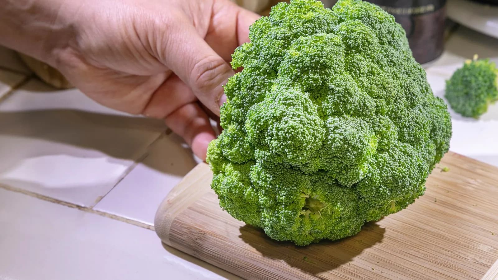 A head of broccoli on a wooden board with a hand steadying it, the thick main stalk still attached below the florets