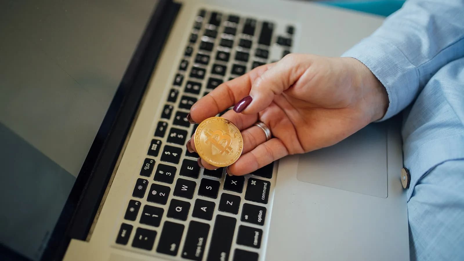 A physical bitcoin token held between finger and thumb in front of an open laptop