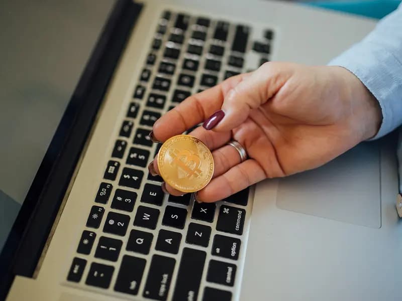 A physical bitcoin token held between finger and thumb in front of an open laptop