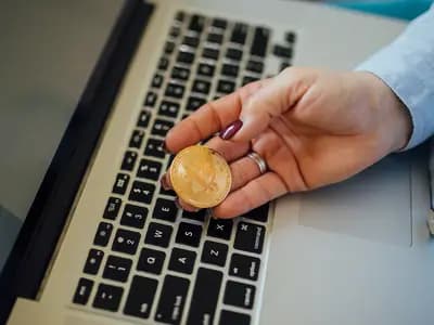 A hand holding a gold coloured metal token stamped with the bitcoin logo above a laptop keyboard. Bitcoin has no physical form and tokens like this one are souvenirs rather than currency