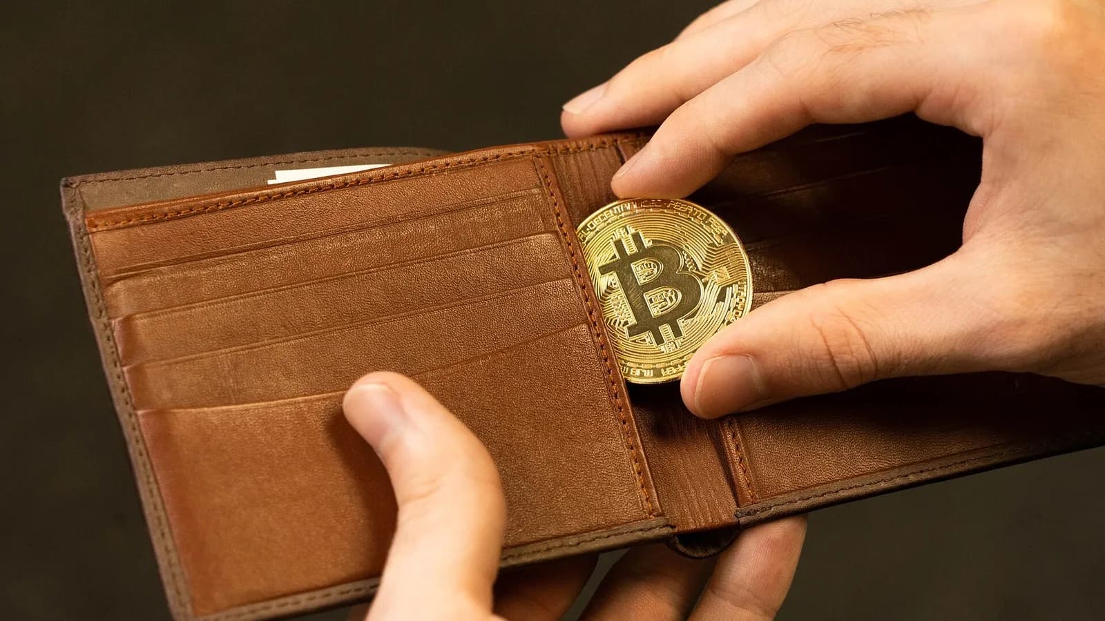 A hand slipping a gold-coloured bitcoin medallion into a brown leather wallet, photographed on 30 July 2021, a stock photograph illustrative of bitcoin and not of any market event