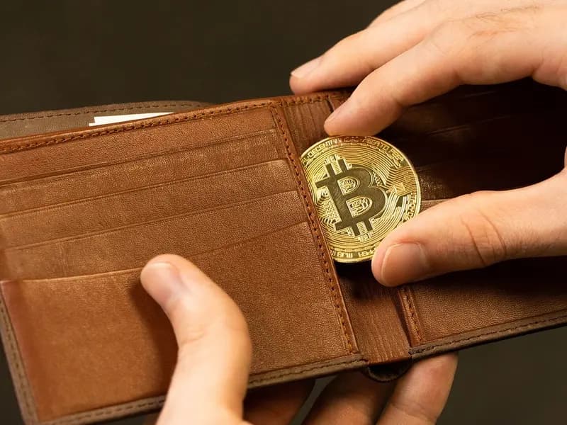 A hand slipping a gold-coloured bitcoin medallion into a brown leather wallet, photographed on 30 July 2021, a stock photograph illustrative of bitcoin and not of any market event