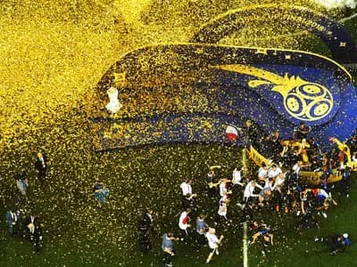 Football players in blue kits celebrating with a trophy on a stadium pitch surrounded by confetti