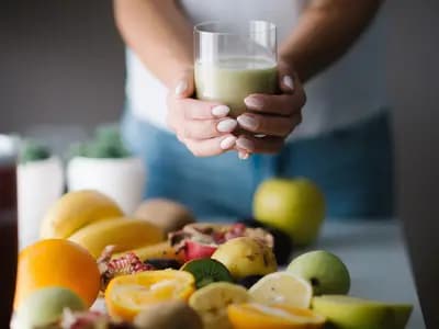 A person holding a glass of green smoothie behind a table of cut fruit