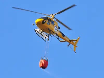 A firefighting helicopter dropping water near a smoke-covered European forest and town