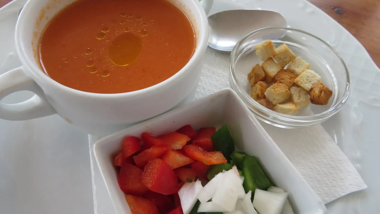 A cup of gazpacho andaluz with a thread of olive oil on top, beside small dishes of diced pepper, cucumber, onion and croutons for garnish