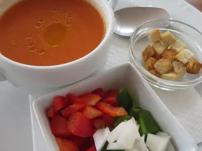 A cup of gazpacho andaluz with a thread of olive oil on top, beside small dishes of diced pepper, cucumber, onion and croutons for garnish