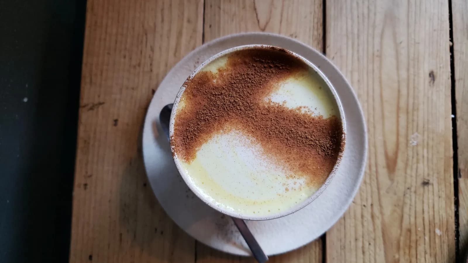 A cup of turmeric milk seen from above on a wooden table, dusted with ground spice across the foam