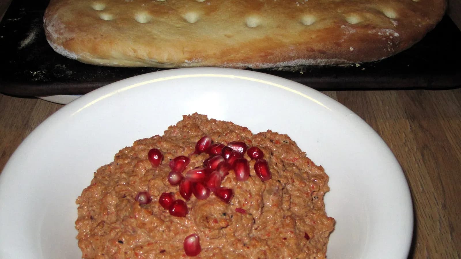 A plate of muhammara scattered with pomegranate seeds, with flatbread behind