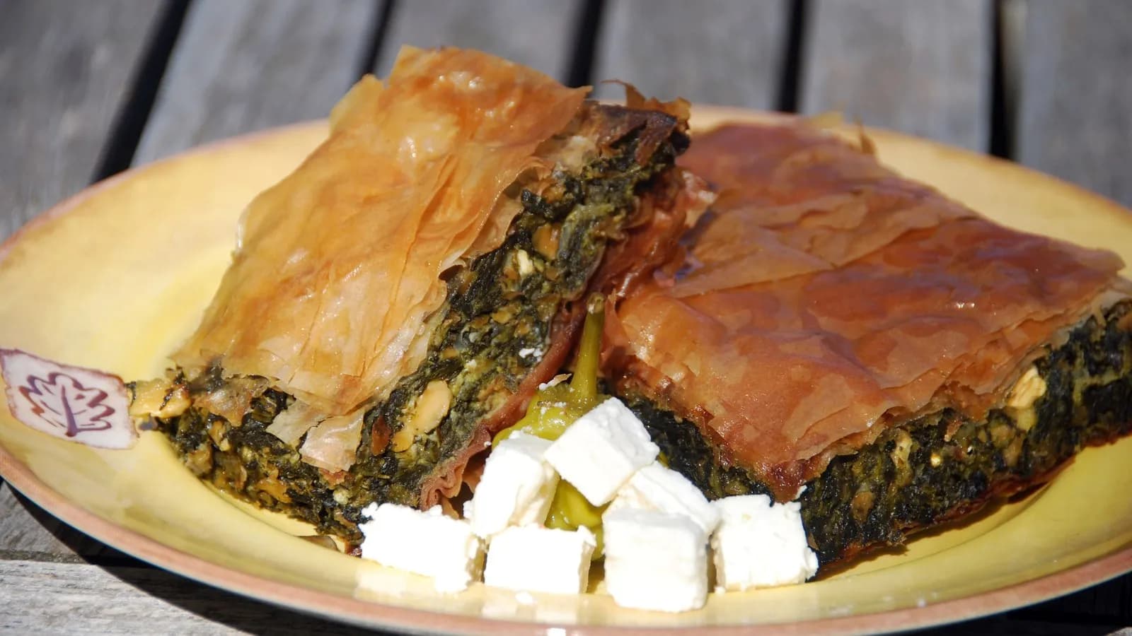 Two wedges of spanakopita on a plate, the layered filo crisp and golden over a dark green spinach filling, with cubes of feta beside them
