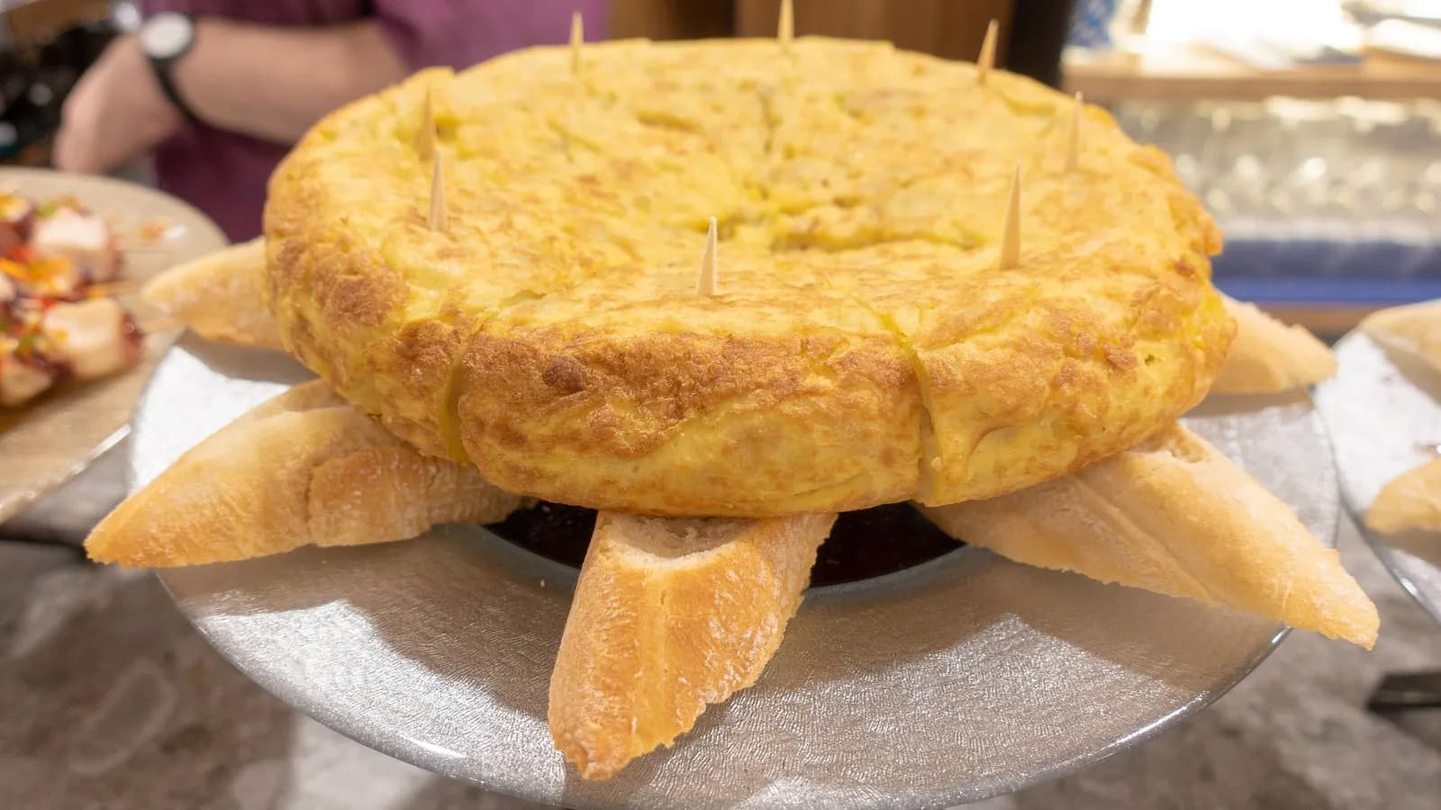 A whole tortilla de patatas on a bar counter in San Sebastián, deep gold and thick, with cocktail sticks and slices of bread around it