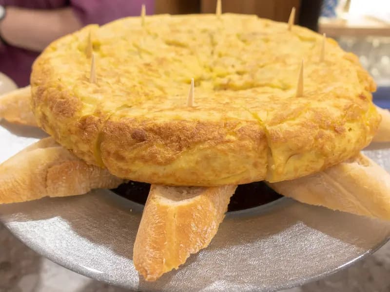 A whole tortilla de patatas on a bar counter in San Sebastián, deep gold and thick, with cocktail sticks and slices of bread around it