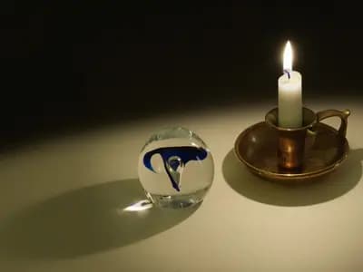 A lit candle in a brass holder on a dark table, its flame refracted through a glass orb beside it